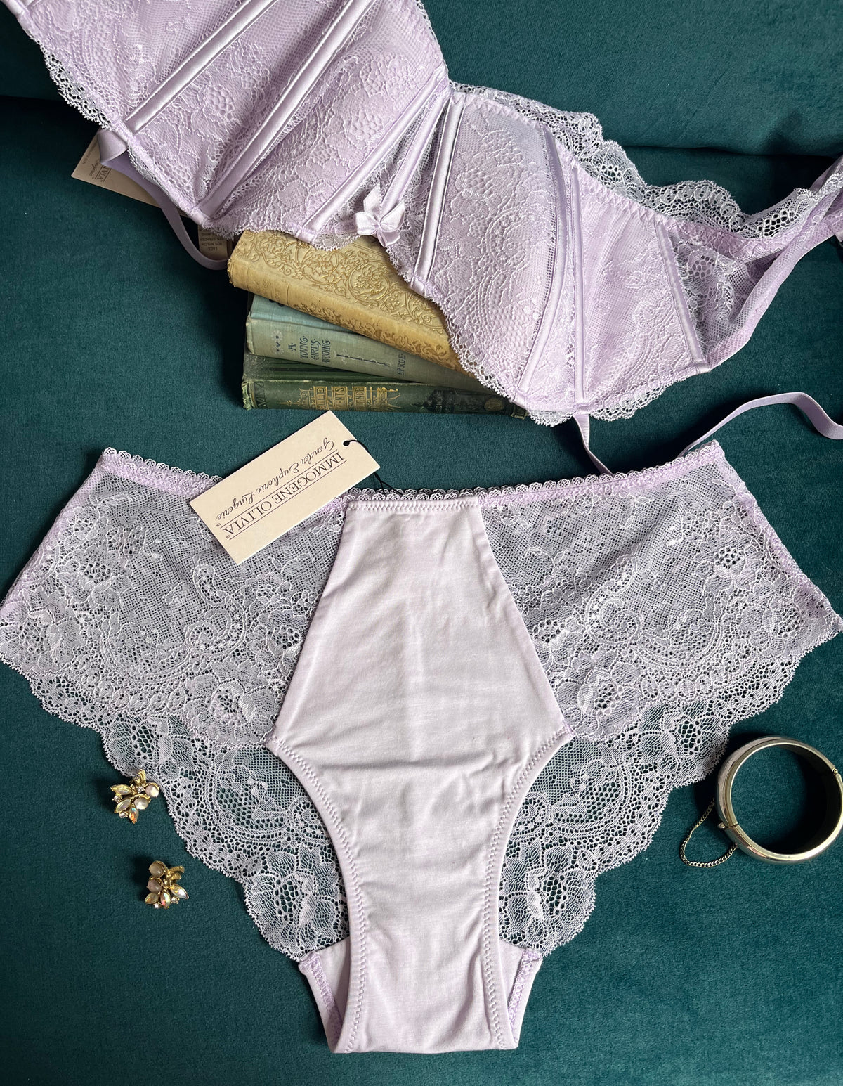Light Purple Lace Camille Lilac Bralette and matching Lace Camille Lilac Panty lace laid flat. Tag shows Immogene Olivia Gender Euphoric Lingerie. Immogene Olivia Gender euphoric lingerie (bra, bralatte, 40 inch band bra, 42 inch band bra, 44 inch band bra, 46 inch band bra, 48 inch band bra, non  tucking panty, mtf, transgender lingerie, gender inclusive, trans women, trans underwear, gender affirming lingerie)