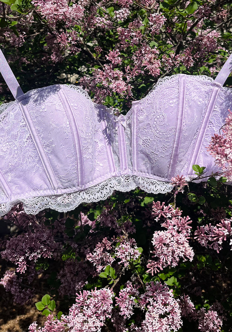 A light purple lace bra from Immogene Olicia Gender Euphoric Lingerie with boning scalloped lace trim, displayed against a background of blooming lilac flowers. (bra, bralatte, 40 inch band bra, 42 inch band bra, 44 inch band bra, 46 inch band bra, 48 inch band bra,  mtf, transgender lingerie, gender inclusive, trans women, trans underwear, gender affirming lingerie)